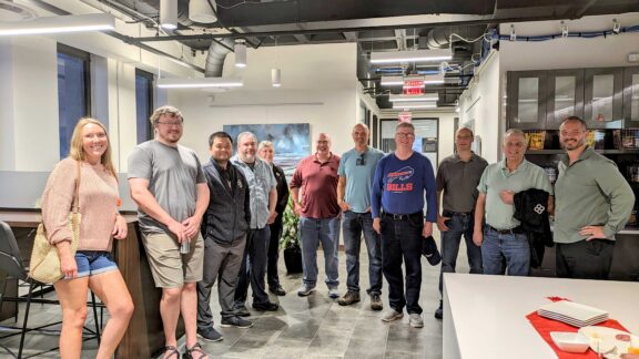 A group of twelve people, eleven men and one woman, stand and smile together in a modern Serendipity Labs Coworking space with white walls, bright lighting, and an open kitchen area near the offices and meeting rooms in the background.