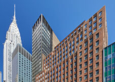 Several tall buildings in New York City, including the art deco-style Chrysler Building on the left, rise against a clear blue sky. Offices and meeting rooms fill many towers, while an American flag and street lamps are visible in the foreground.