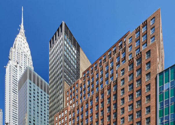 Several tall buildings in New York City, including the art deco-style Chrysler Building on the left, rise against a clear blue sky. Offices and meeting rooms fill many towers, while an American flag and street lamps are visible in the foreground.