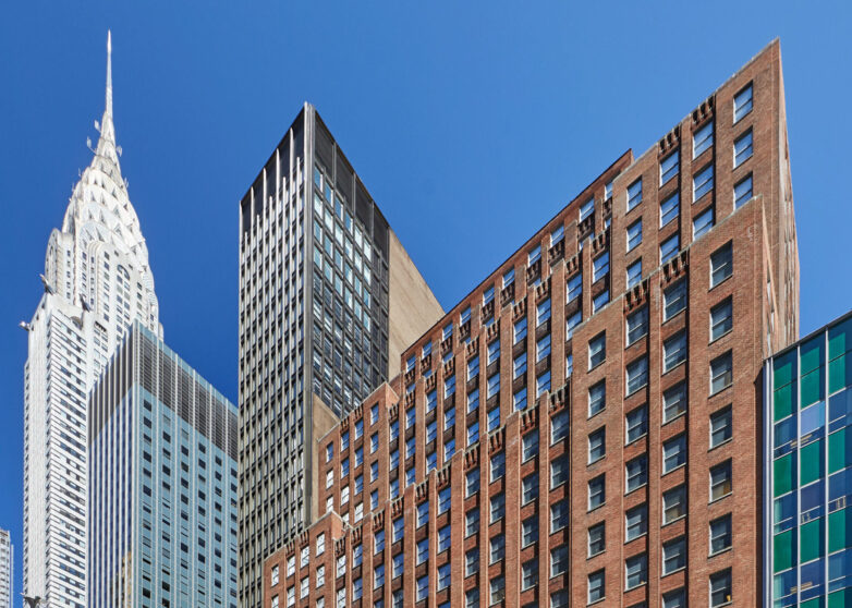 Several tall buildings in New York City, including the art deco-style Chrysler Building on the left, rise against a clear blue sky. Offices and meeting rooms fill many towers, while an American flag and street lamps are visible in the foreground.