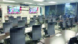 A modern classroom with rows of empty desks and gray chairs facing two large screens, reminiscent of offices and meeting rooms at Serendipity Labs Coworking. Large windows fill the space with natural light and offer a view of trees outside.