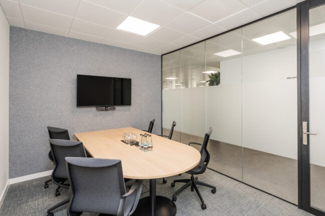A modern conference room at Serendipity Labs Coworking features a wooden oval table, six gray office chairs, a wall-mounted TV, glass walls, and bottles of water—perfect for productive meetings in well-lit offices and meeting rooms.
