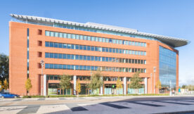 Modern six-story office building with large glass windows and red brick exterior, set against a clear blue sky. Trees and a few parked cars are in front, with offices and meeting rooms available and a To Let sign on the ground floor.