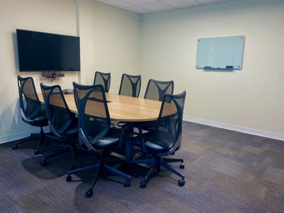 A modern conference room at Serendipity Labs Coworking features a wooden oval table, eight black mesh office chairs, a wall-mounted TV, and a whiteboard on a beige wall—perfect for offices and meeting rooms needs.