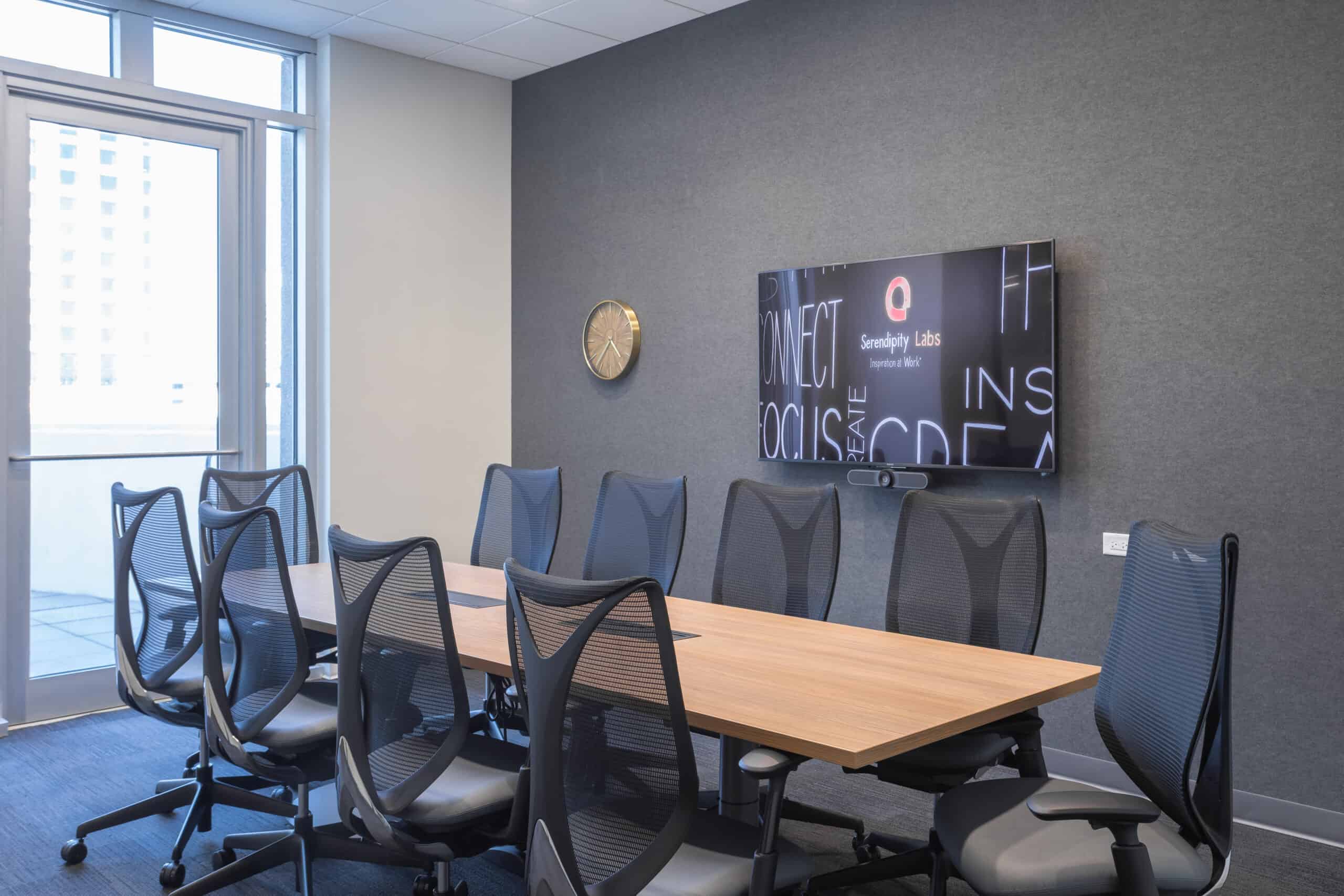 A modern conference room with a long wooden table, ten mesh office chairs, a wall-mounted TV displaying text, a round wall clock, and large windows letting in natural light.