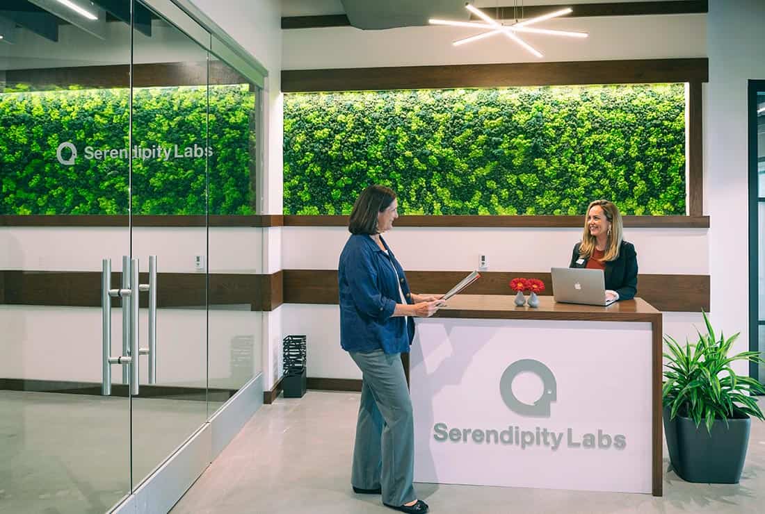 Two women converse at the Serendipity Labs coworking reception desk in a modern office with a green moss wall, glass doors, and potted plants—showcasing stylish offices and meeting rooms. One stands holding a folder while the other smiles behind the desk.