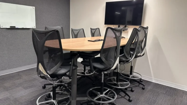 A modern conference room at Serendipity Labs coworking features a wooden oval table, seven high mesh chairs, a wall-mounted TV, and a small whiteboard on a gray wall—with dark gray carpet flooring. Ideal for offices and meeting rooms.