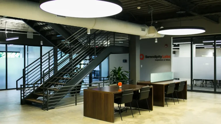 Modern office space with wooden desks and black chairs, large overhead lights, a metal staircase, glass-walled offices and meeting rooms, and a Serendipity Labs sign on the wall. The serendipity labs coworking area looks clean and organized.