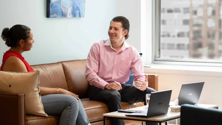 Two people sit on a brown couch in a bright serendipity labs coworking space, engaged in conversation. Both have laptops open on the table, with city buildings visible through the large windows of these modern offices and meeting rooms.
