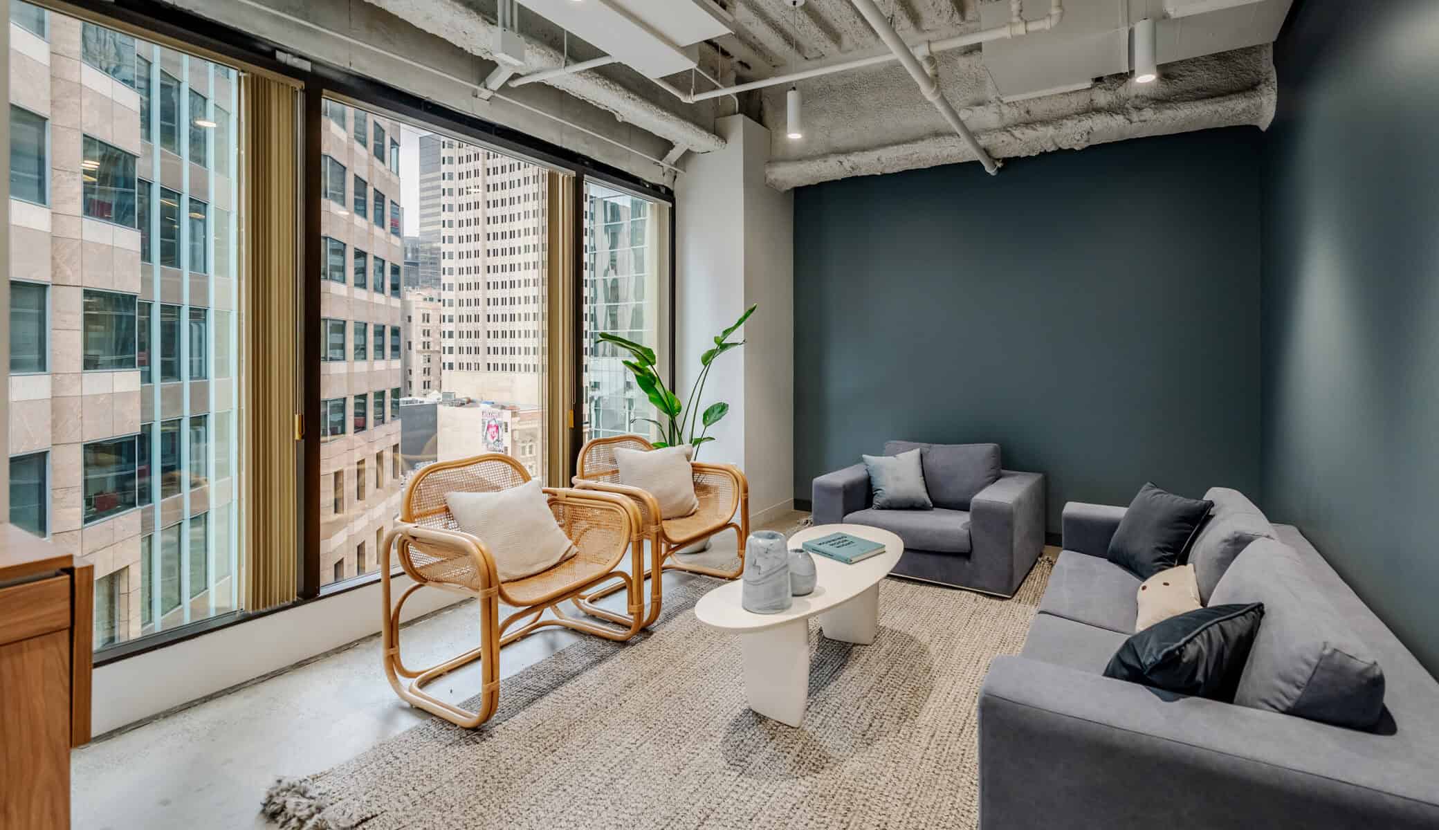 Modern office lounge ideal for film shoots or photography shoots, featuring large windows, gray sofas, wicker chairs, a white coffee table, and potted plant. City buildings are visible outside, with exposed ceilings and a neutral color palette.