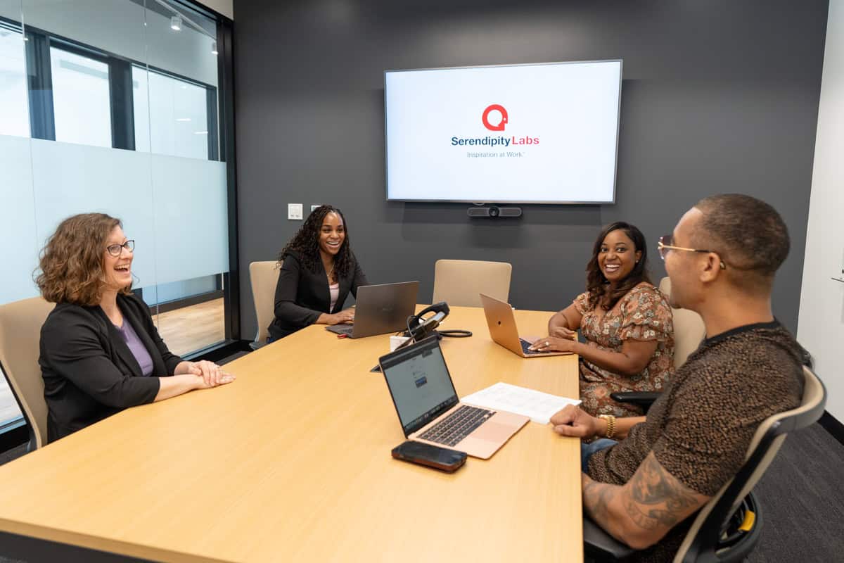 Four people sit around a conference table with laptops, smiling and having a meeting in a Serendipity Labs coworking space. A screen displays the logo, highlighting the professional and collaborative atmosphere of their offices and meeting rooms.