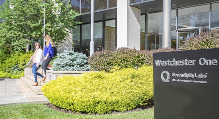 Two women walk down steps outside a modern office building with large windows. In the foreground, a sign reads Westchester One and Serendipity Labs Coworking, highlighting its offices and meeting rooms amid lush greenery.White Plains Welcome serendipity labs