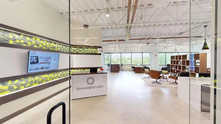 Modern office space with a reception desk labeled “Serendipity Labs,” green moss wall accents, lounge chairs, bookshelves, and large windows. The open ceiling features exposed beams and lights in this vibrant serendipity labs coworking hub with offices and meeting rooms.