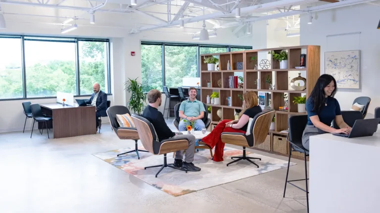 Modern office space with large windows, featuring serendipity labs coworking—four people chat in lounge chairs around a coffee table, one works on a laptop at a counter, and another at a desk near the window as natural light fills the offices and meeting rooms.