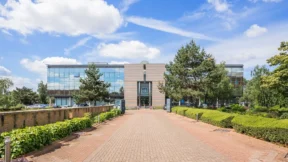 A modern office building with large glass windows, featuring offices and meeting rooms, is surrounded by greenery and trees, viewed from a brick walkway under a blue sky with scattered clouds.