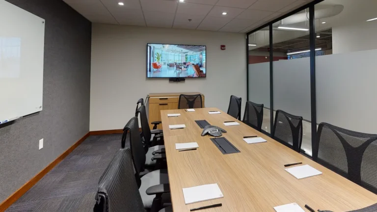 A modern conference room at Serendipity Labs Coworking, featuring a long wooden table, ten black mesh chairs, notepads with pens, a wall-mounted screen showing offices and meeting rooms, a whiteboard, and glass walls with frosted panels.