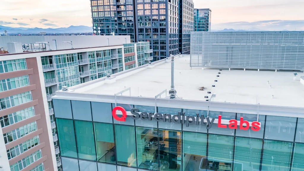 A modern glass building with Serendipity Labs signage on its side, offering offices and meeting rooms, stands amid other office and apartment buildings, with mountains visible in the distant background.