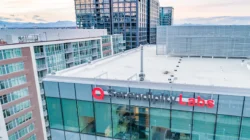 A modern glass building with Serendipity Labs signage on its side, offering offices and meeting rooms, stands amid other office and apartment buildings, with mountains visible in the distant background.