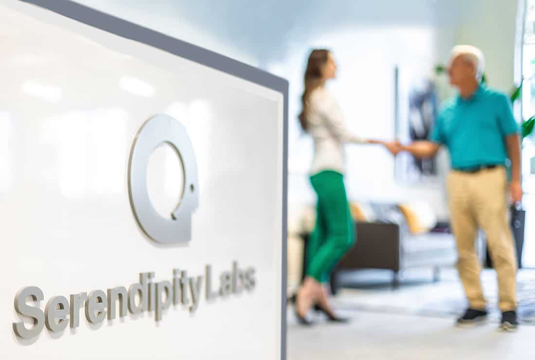 Two individuals shaking shades behind of the Serendipity Labs logo