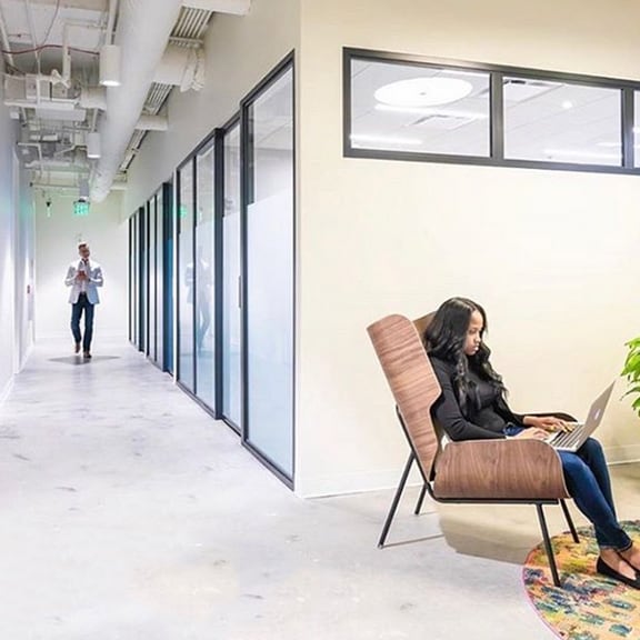 A woman independently in Coworking area, while a man walks past the private office rooms.