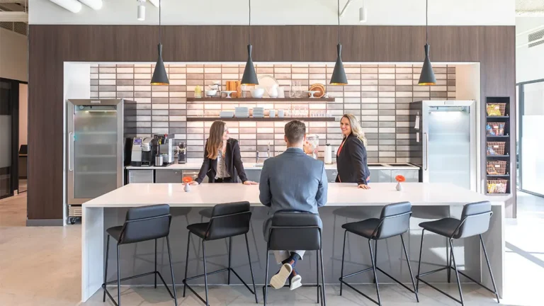 Three people in business attire talk around a modern office kitchen island at Serendipity Labs coworking. Shelves, appliances, and pendant lights decorate the background in this bright, contemporary space with offices and meeting rooms.