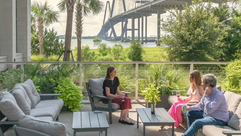 Three people sit and talk on a patio with cushioned furniture, surrounded by greenery. In the background, offices and meeting rooms overlook a river and a large cable-stayed bridge under a partly cloudy sky.