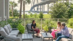 Three people sit and talk on a patio with cushioned furniture, surrounded by greenery. In the background, offices and meeting rooms overlook a river and a large cable-stayed bridge under a partly cloudy sky.