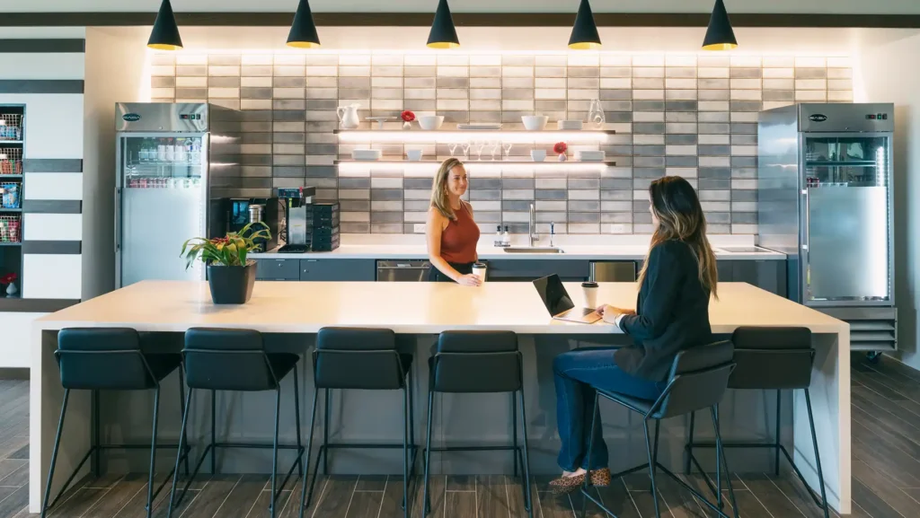Two women are in a modern Serendipity Labs coworking office kitchen with a long countertop, bar stools, and hanging lights. One stands by the sink while the other sits with a laptop and coffee. Shelves, fridges, and a potted plant are visible.