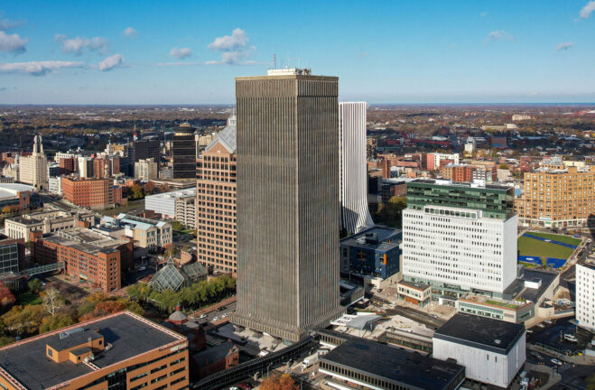 Aerial view of a city skyline featuring a tall, rectangular skyscraper at the center, surrounded by various office buildings with modern offices and meeting rooms, including Serendipity Labs coworking spaces beneath a partly cloudy sky.