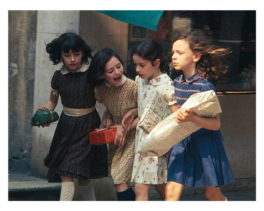 Four young girls walk together outdoors, holding wrapped gifts and packages, with joyful and animated expressions. They are dressed in vintage-style dresses and appear to be in mid-conversation or laughter.