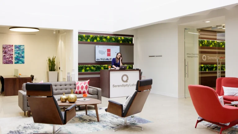 Modern office lobby with colorful seating, a gold coffee table, and green plant wall. A woman stands at the Serendipity Labs coworking reception desk; glass offices and meeting rooms plus artwork are visible in the bright, open space.