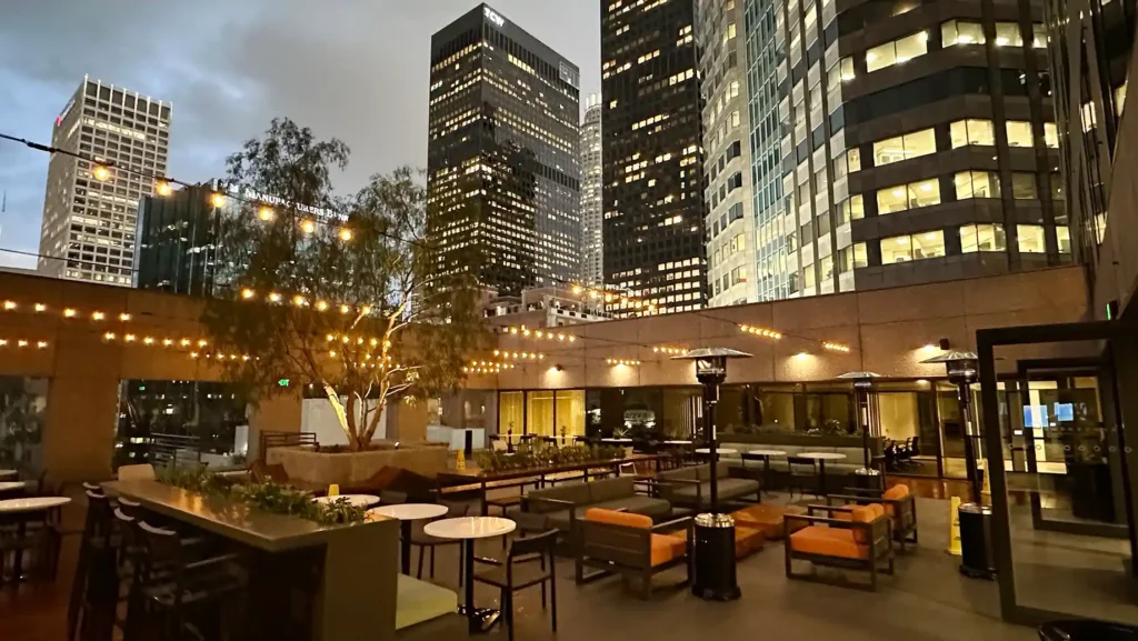 A rooftop patio at dusk features string lights, outdoor sofas, and tables, surrounded by skyscrapers with illuminated windows—perfect for unwinding after a day at Serendipity Labs coworking offices and meeting rooms in the city.